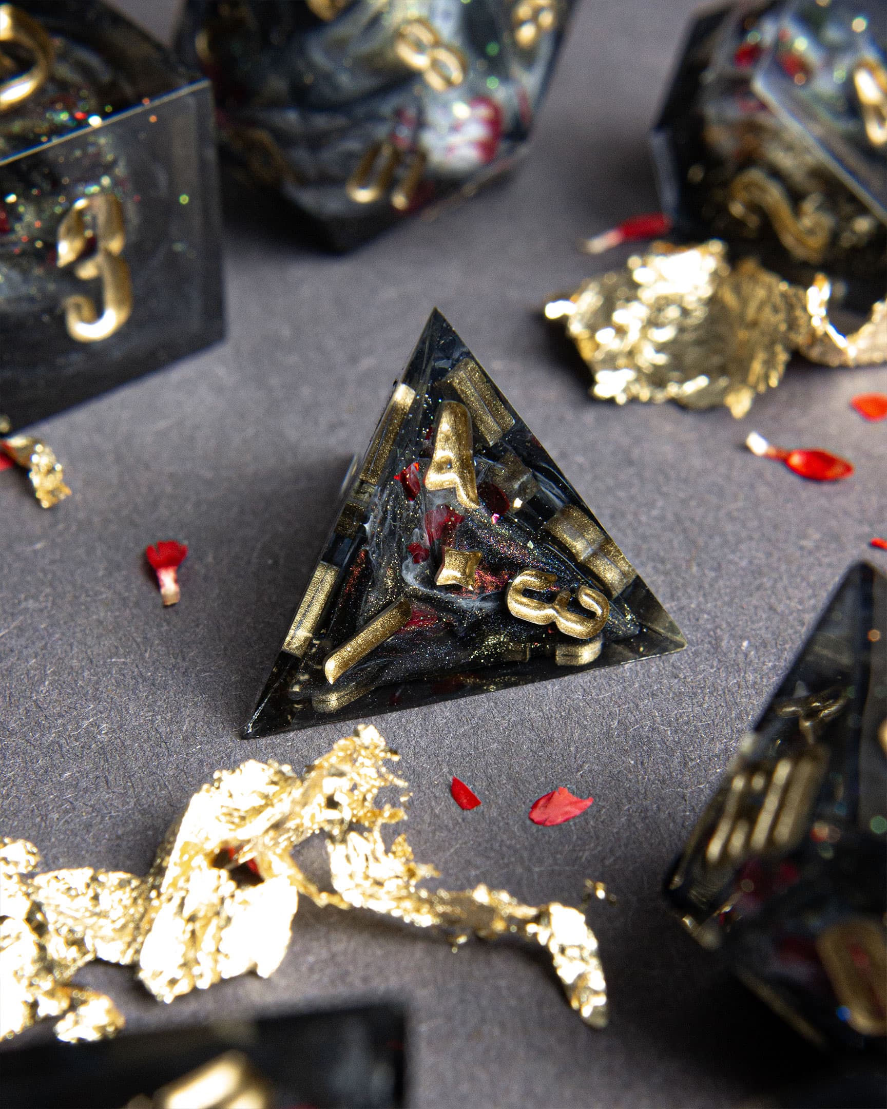 The Last Gommage D4 Pyramid, black resin with gray swirls, red flower petals, metallic foil, and golden numbers